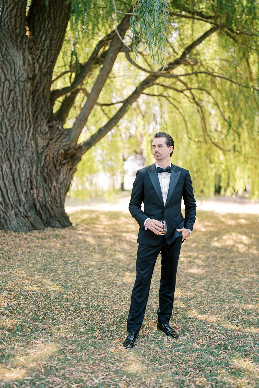 Man in formal black tuxedo holding a glass, standing under a large tree in a sunlit park setting