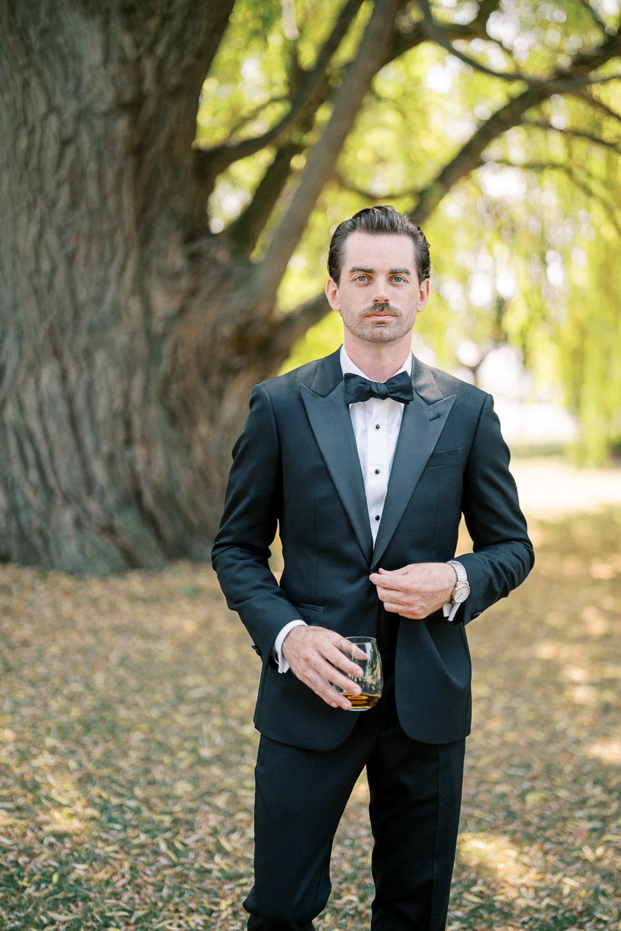 Man in a black tuxedo holding a drink in a sunny outdoor setting with trees in the background.