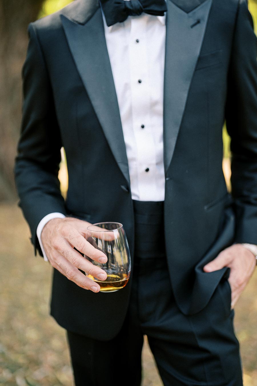 Man in a black tuxedo holding a glass of whiskey outdoors.