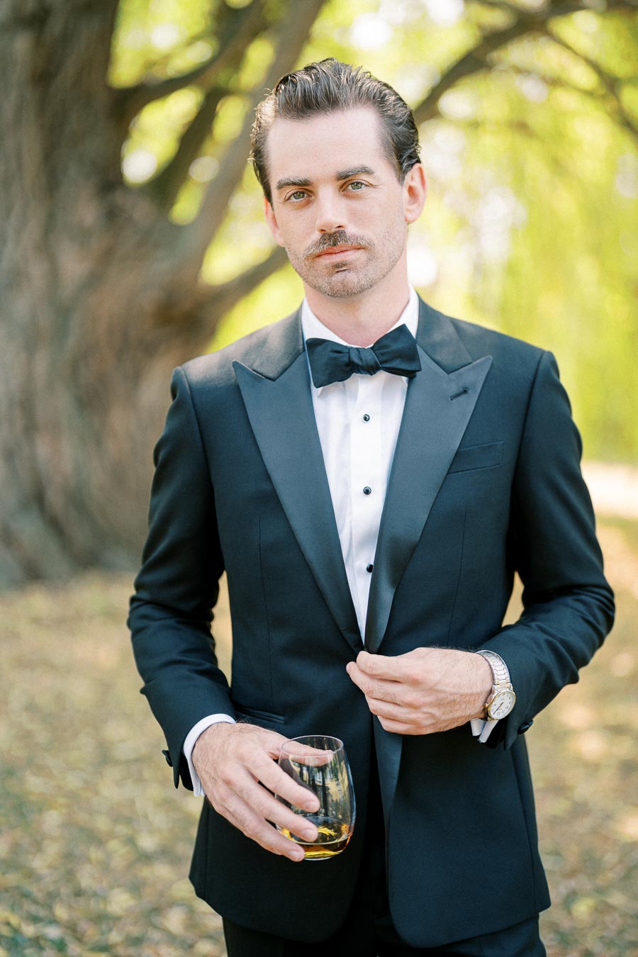 A well-dressed man in a classic black tuxedo holding a glass of whiskey outdoors, standing confidently beneath a tree with blurred greenery in the background.
