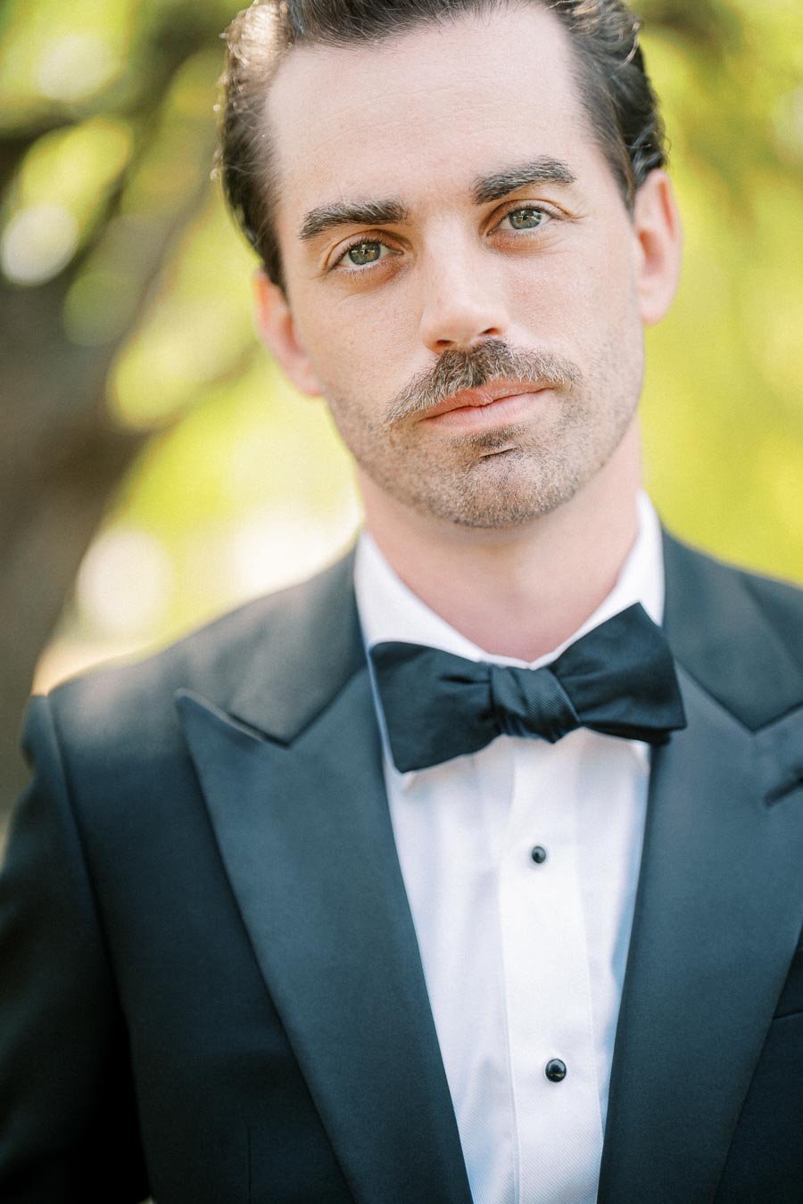 A man in a formal black tuxedo with a bow tie standing outdoors with a blurred natural background.