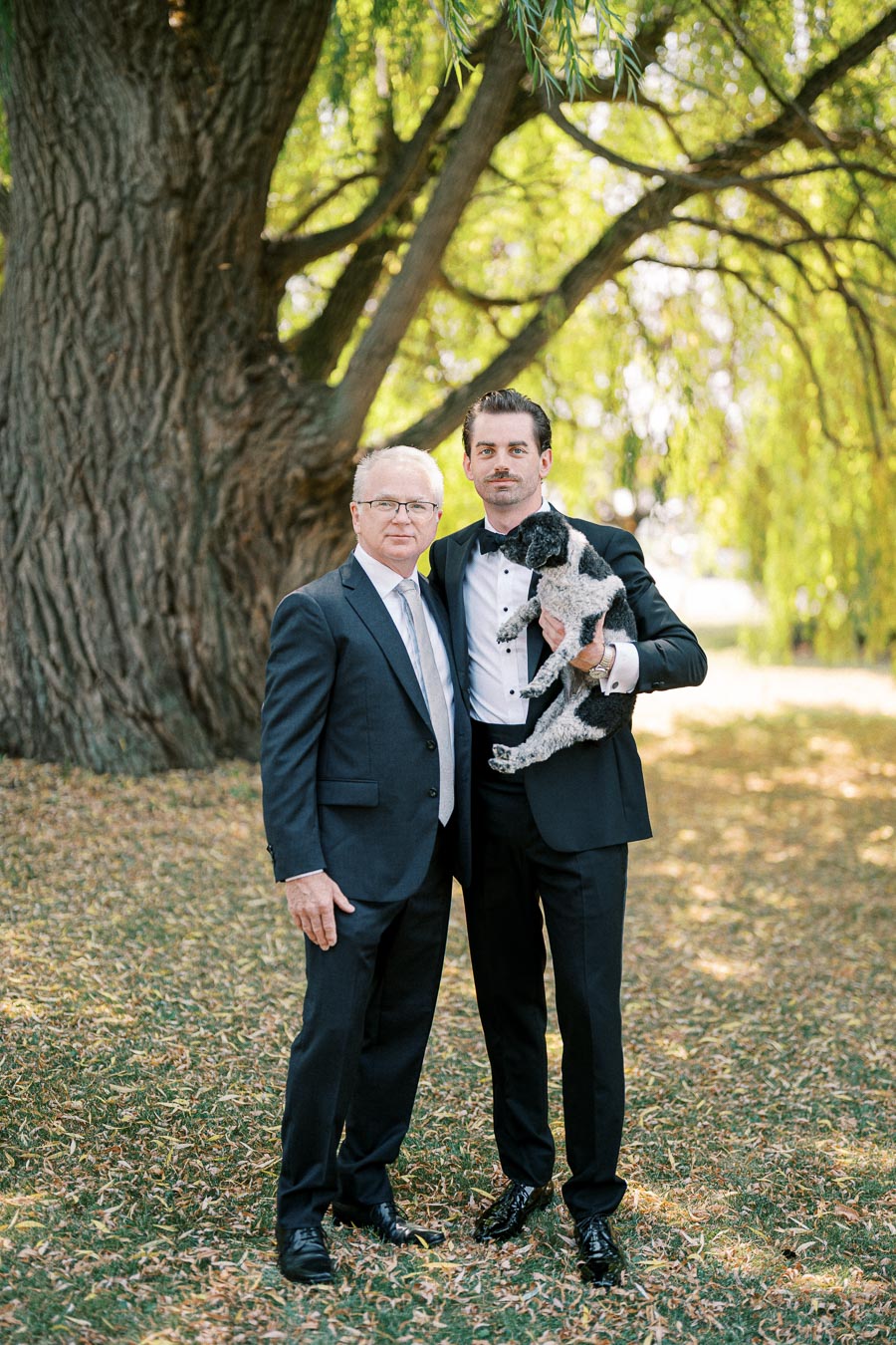 Two men in formal suits standing under a large tree, one holding a black and white dog, outside in a park setting.