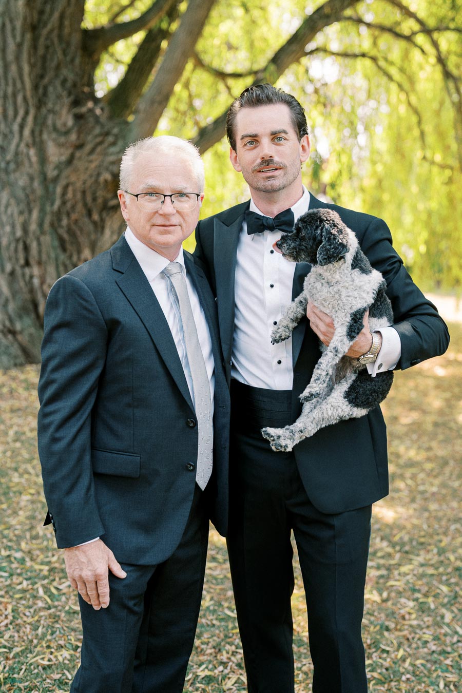 Two men in formal suits standing side by side, with one holding a small black and white dog, outdoors in a park setting with a large tree and green foliage in the background.