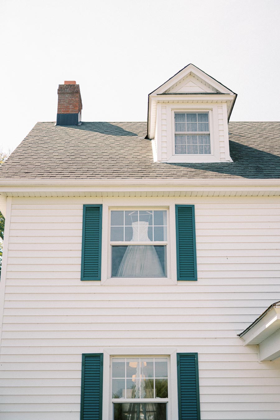 Exterior view of a white house with teal shutters, featuring a bridal gown displayed in the upper window, chimney visible on the left, under a clear sky.