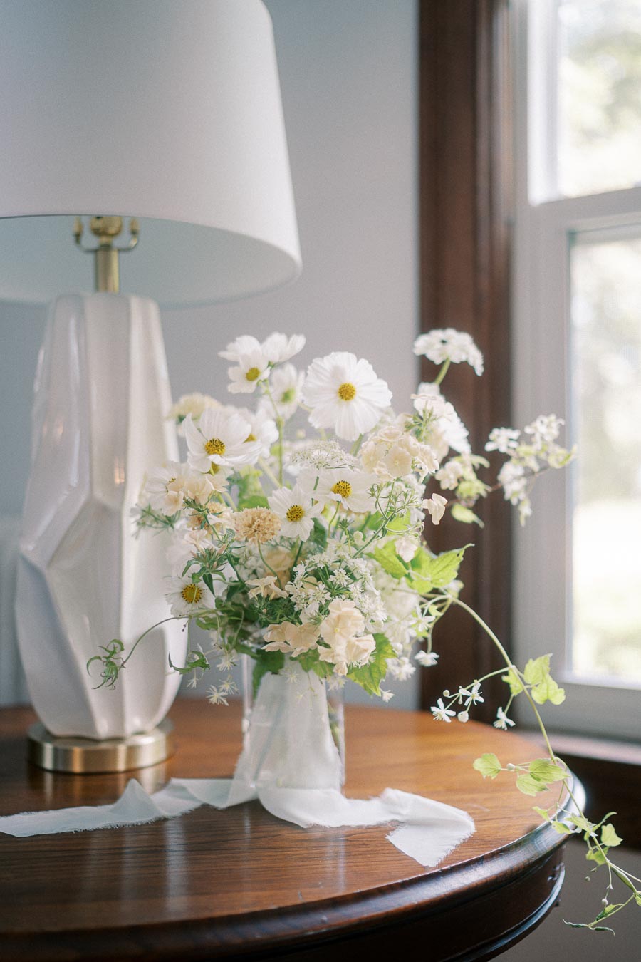 Elegant white floral arrangement in a clear vase on a wooden table, next to a modern white lamp, with natural light from a nearby window.