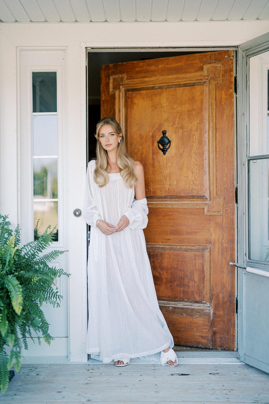 A woman in a flowing white dress stands at a wooden door entrance, exuding elegance and grace. The scene is complemented by a potted fern on a wooden porch, suggesting a peaceful and welcoming atmosphere.