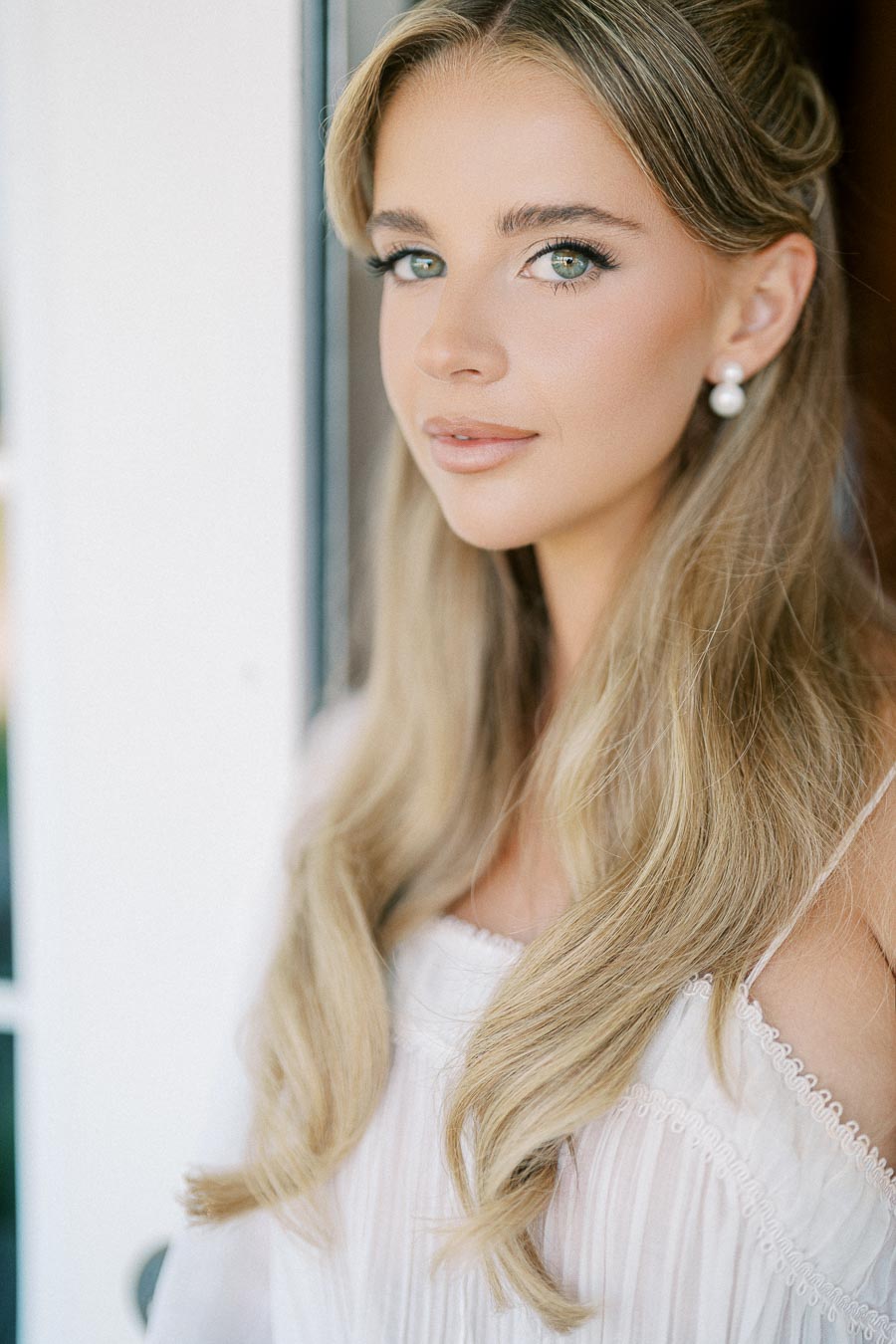 Young woman with long blonde hair and pearl earrings, wearing a white dress, looking directly at the camera with a gentle expression