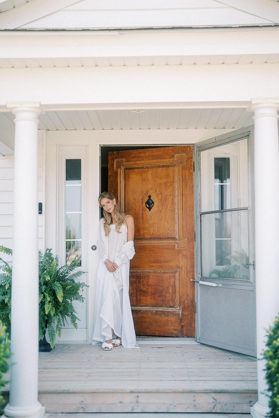 A woman in a flowing white dress standing on a wooden porch next to a large wooden front door, with greenery and white pillars framing the entrance.