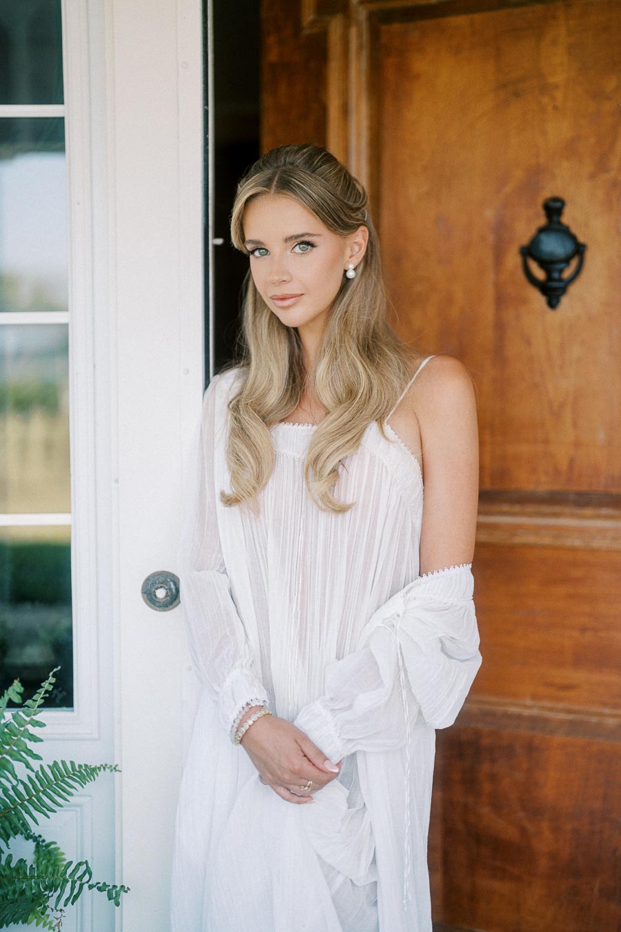 Woman in elegant white dress standing by wooden door, showcasing long wavy hair and pearl earrings, with natural makeup and soft lighting.