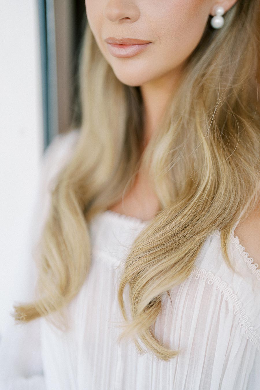 Close-up of a woman's lower face showcasing soft, natural makeup with pearl earrings and long, wavy blonde hair cascading over a white, textured blouse.