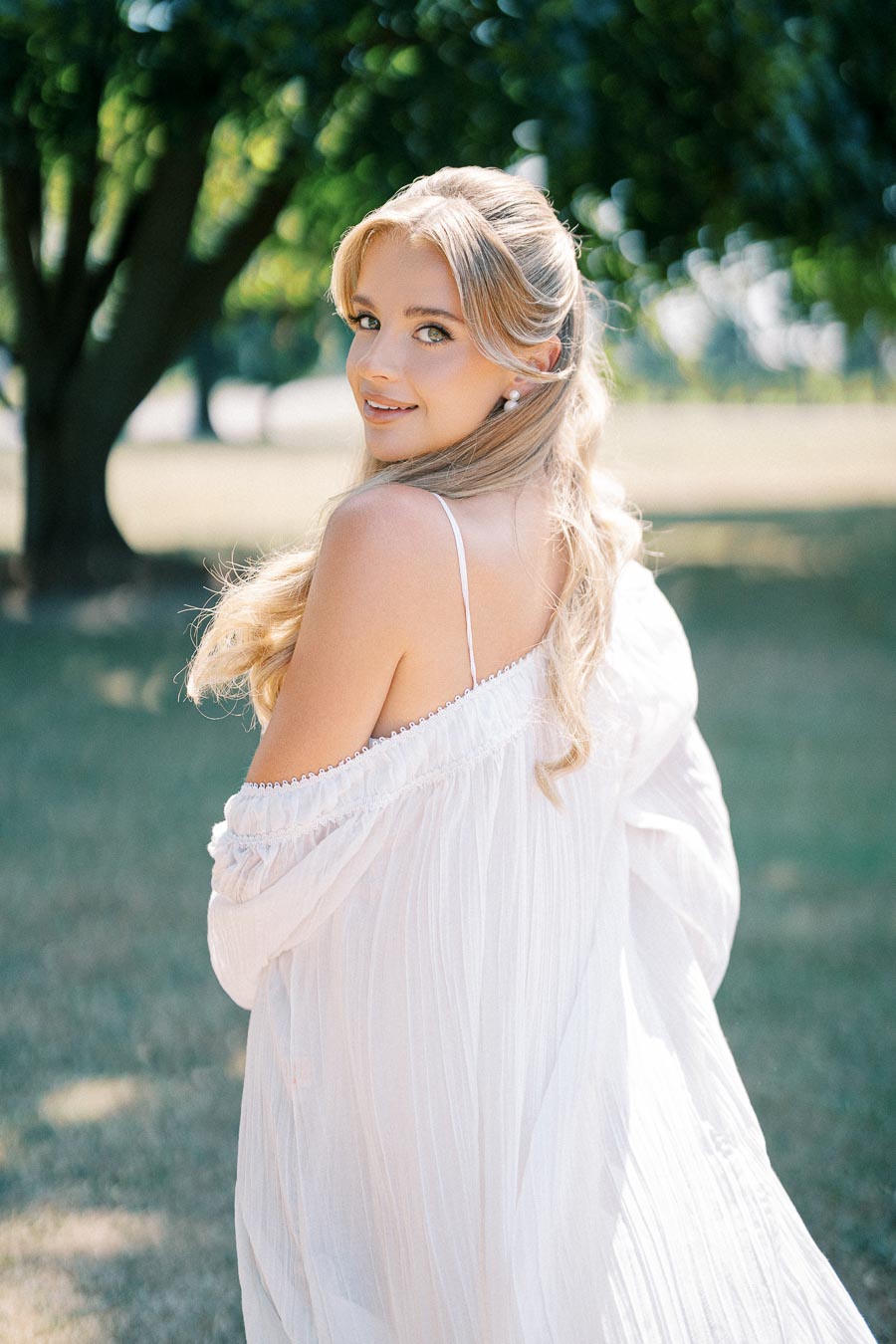 Woman in a flowing white dress smiling in a sunlit park setting, with green trees in the background.
