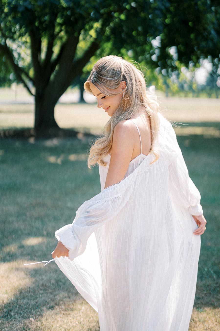 A woman in a flowing white dress gracefully poses outdoors in a sunlit garden, with lush green trees in the background.