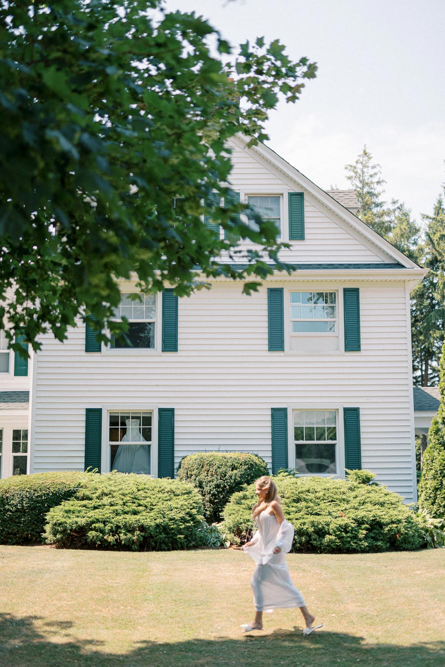 Elegant woman walking on lawn in front of a classic white house with green shutters, surrounded by lush greenery and trees.