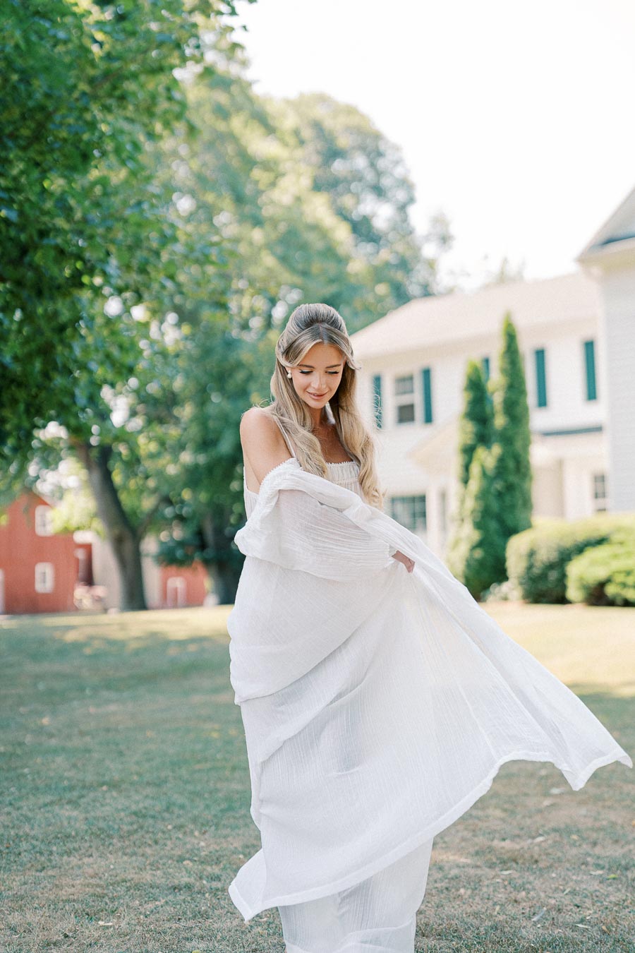 A woman in a flowing white dress stands gracefully on a grassy lawn in front of a large, elegant house, surrounded by lush green trees.