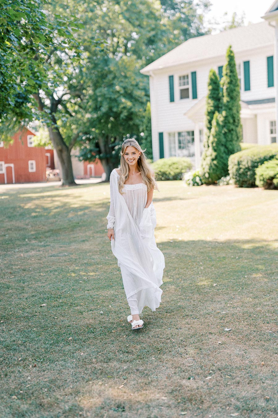 A woman in a flowing white dress walking on a lawn in front of a house, with greenery and a red barn in the background on a sunny day.