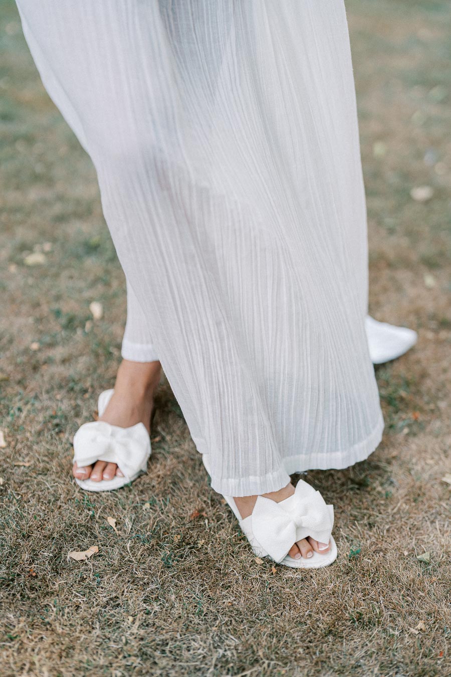 Elegant white sandals with large bows paired with a flowing white dress, worn outdoors on a grassy surface.