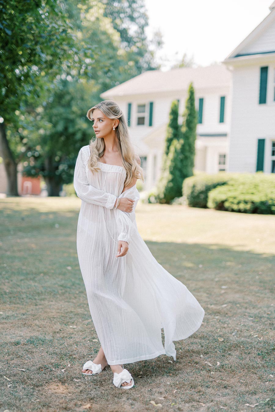 A woman in a long, flowing white dress stands elegantly on a grassy lawn in front of a white house with green shutters, surrounded by lush greenery on a sunny day.