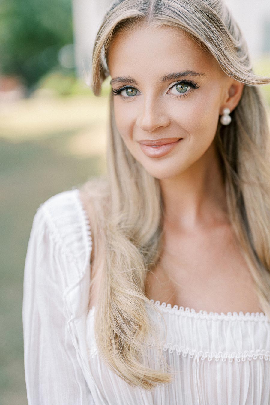 A woman with long blonde hair and pearl earrings smiling outdoors, wearing a white, textured top.