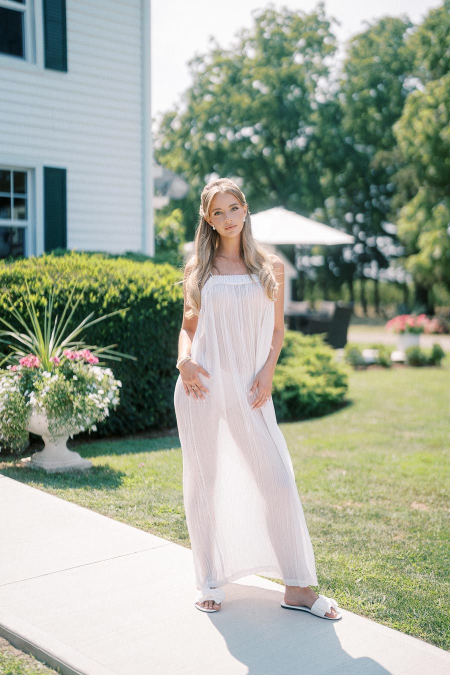 A woman in a flowing white summer dress stands outdoors on a sunny day, in front of a house with vibrant green shrubs and colorful flowers. She is positioned on a sidewalk, with a garden and greenery visible in the background.