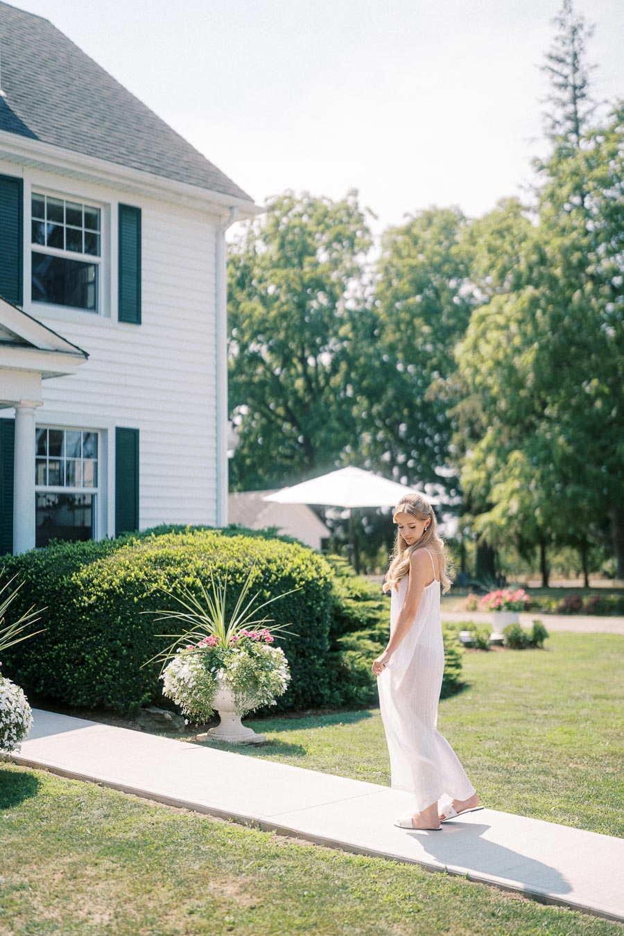 A woman in a flowing white dress walking on a garden path near a traditional white house with green shutters, bordered by vibrant greenery and potted flowers, under a clear blue sky.