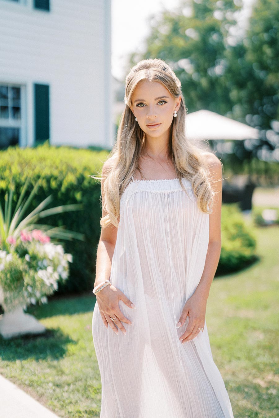A woman in an elegant, flowing white dress stands outdoors in a sunlit garden setting, with blurred greenery and a house in the background, capturing a serene and stylish summer look.