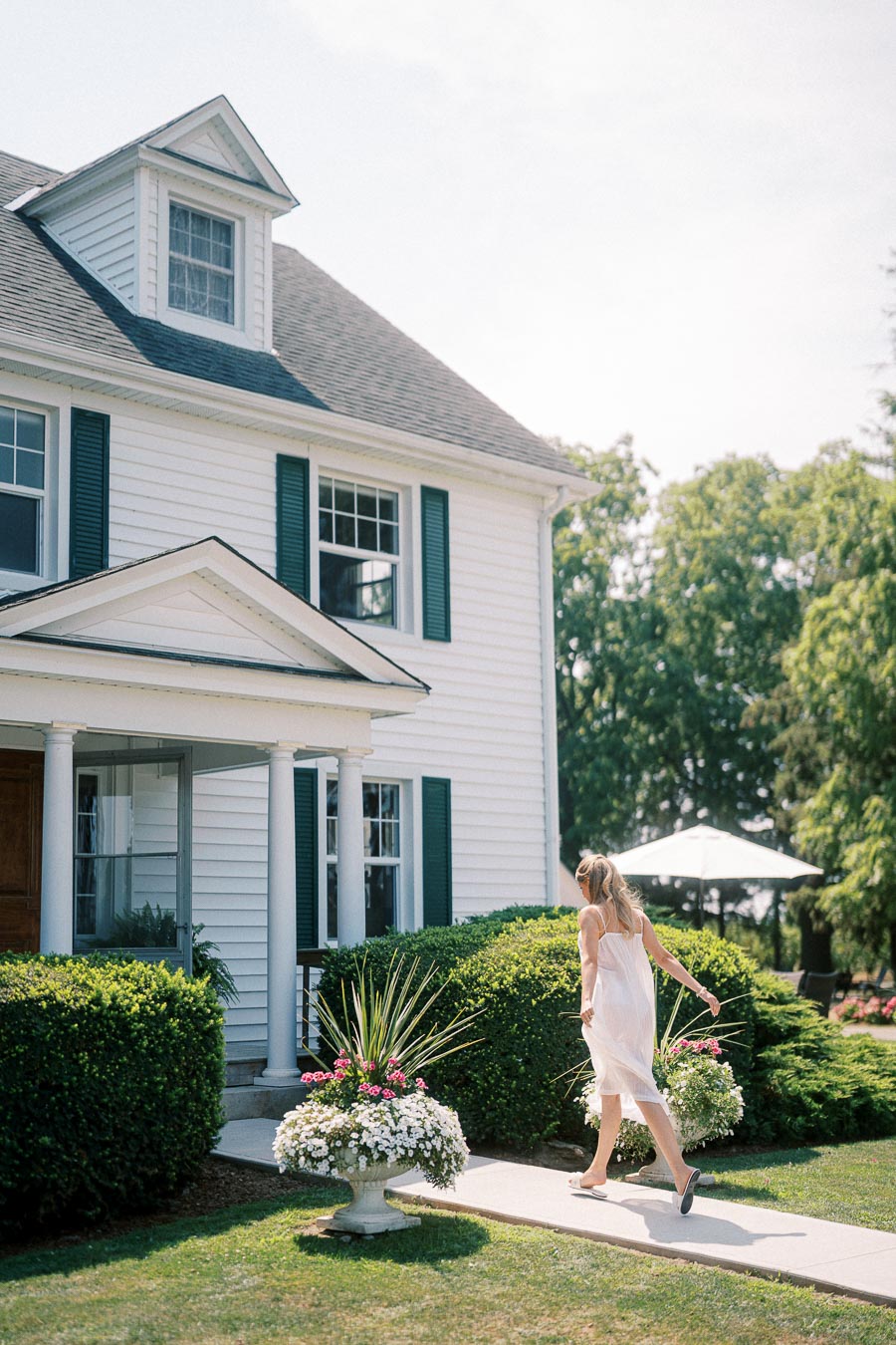 A woman in a white dress walks towards a charming white house with green shutters surrounded by lush greenery and flowers under a sunny sky.