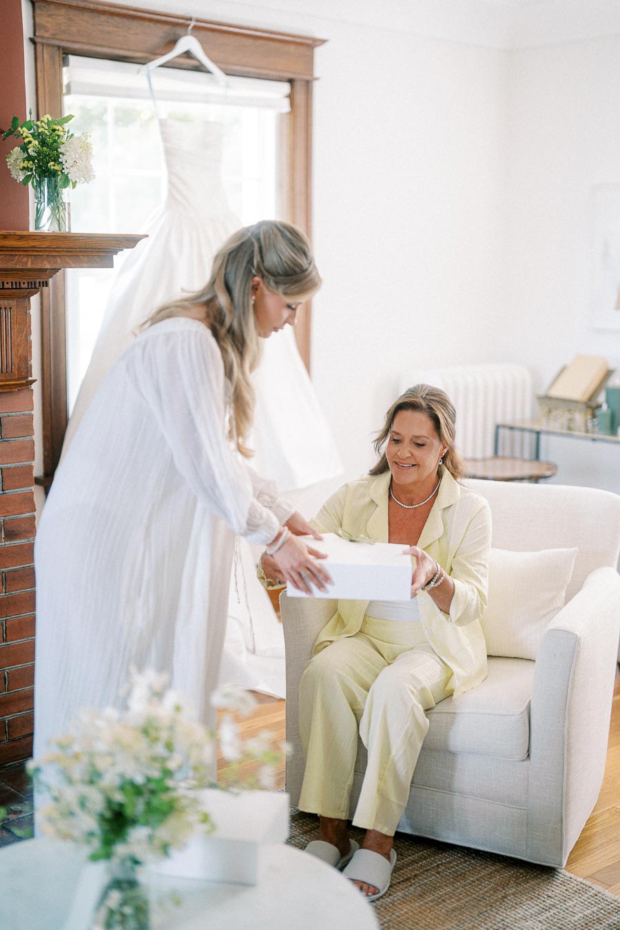 A woman in a white dress hands a gift box to another woman in a yellow outfit seated on a cream chair in a bright room. A wedding dress hangs in the background near a fireplace with flowers, suggesting a bridal preparation scene.