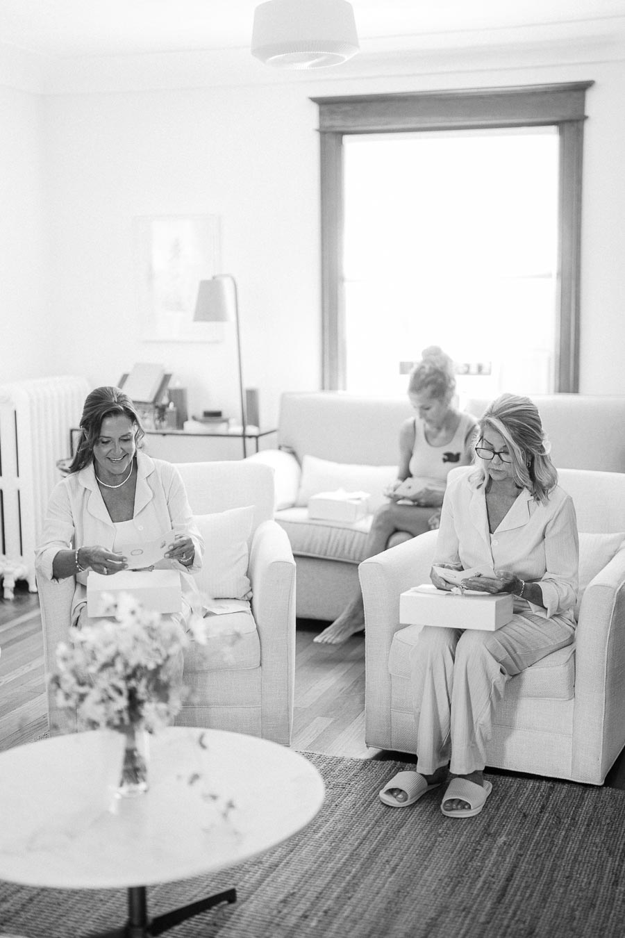 Black and white image of women sitting in a cozy living room while opening gifts. They appear relaxed, wearing comfortable clothing, with a vase of flowers on a round table in the foreground. Bright natural light fills the room through the window, creating a warm and inviting atmosphere.