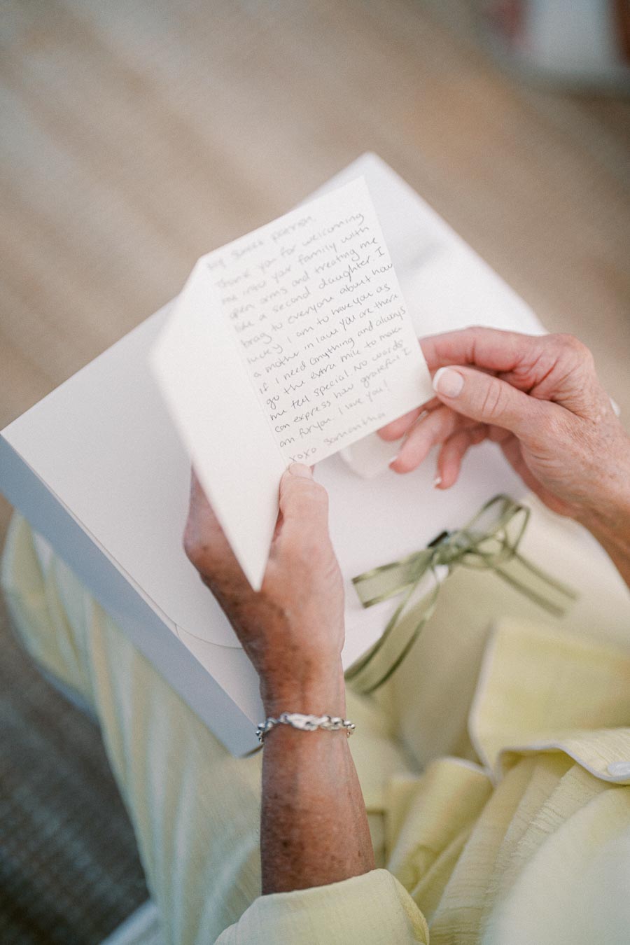 Elderly person reading a heartfelt handwritten letter, seated in a light fabric outfit, next to a gift box with a green ribbon.