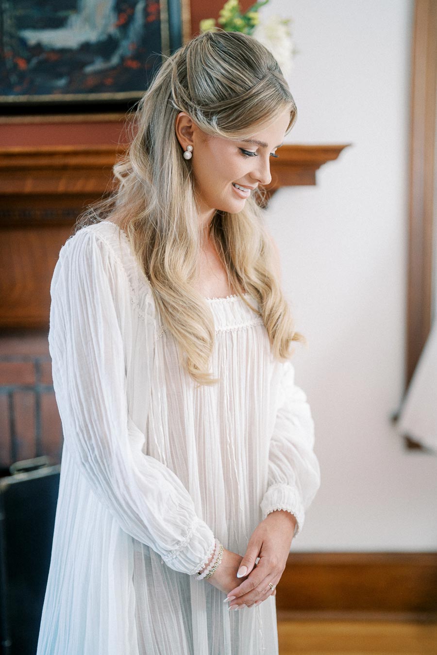 Smiling woman in elegant white dress with long, wavy blonde hair, standing indoors near a wooden mantelpiece.