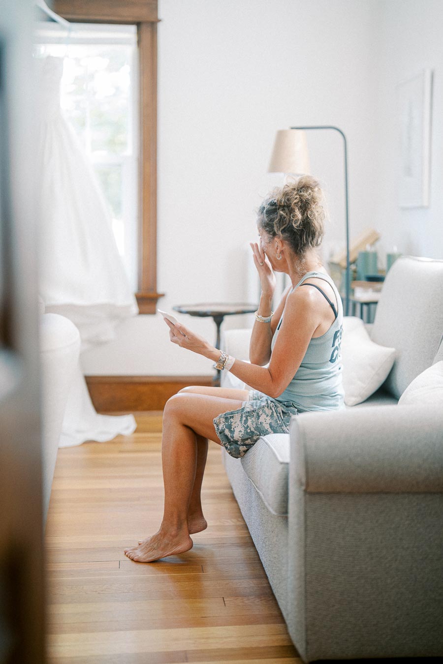 Woman sitting on a couch in a softly lit room, wiping tears while looking toward a white dress hanging near a window, evoking emotions of joy and nostalgia.