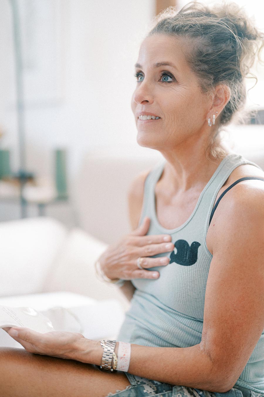 Middle-aged woman with an expressive look, wearing a light blue tank top with a bird design, sitting on a white couch, hand placed on her chest, engaged in reading a handwritten letter, conveying emotion and thoughtfulness in a bright, cozy interior.