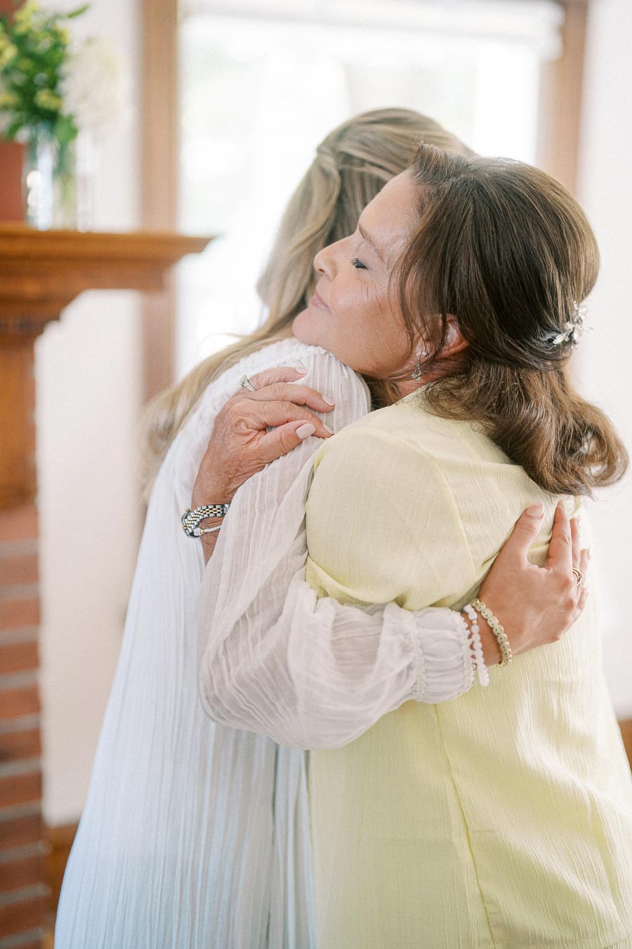 Two women embracing warmly in a cozy room, one wearing a light-colored dress and the other in a pastel yellow outfit with floral hair accessories, expressing affection and connection.