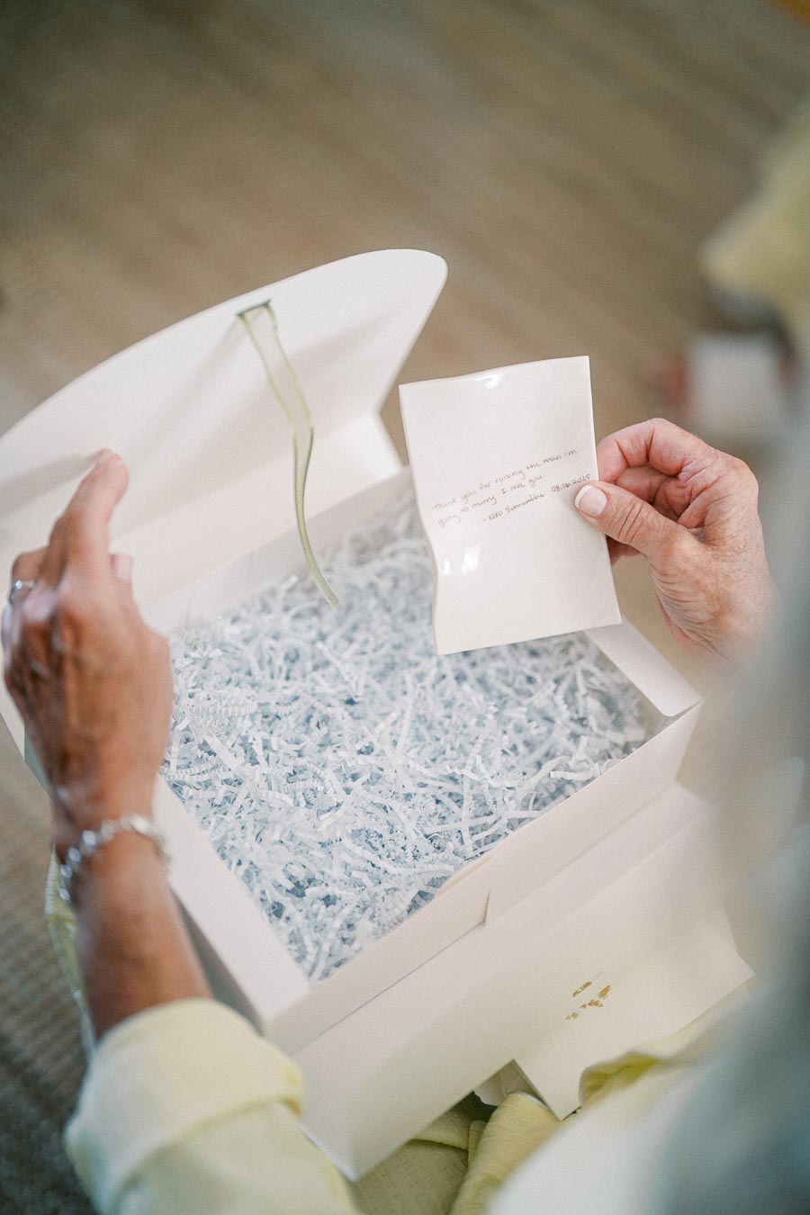 An older person opens a white gift box filled with shredded paper, holding a handwritten note.