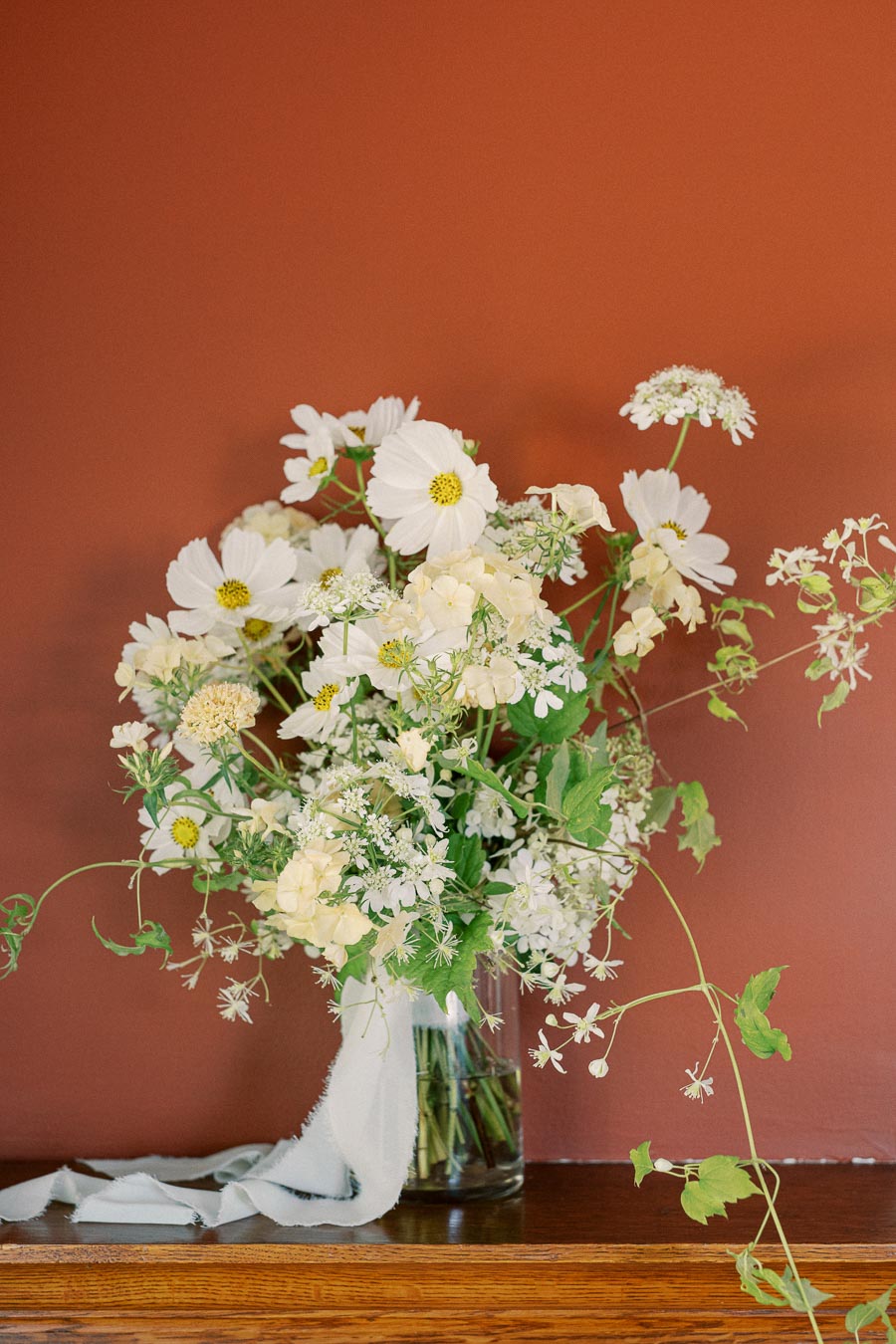 A vase of delicate white wildflowers including daisies and hydrangeas set against a warm terracotta-colored wall, with a soft fabric draped around the arrangement.