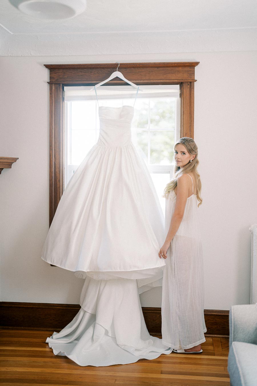 A bride stands next to a hanging white wedding gown in a bright room, showcasing an elegant window backdrop and wooden trim, exuding a serene and classic bridal atmosphere.