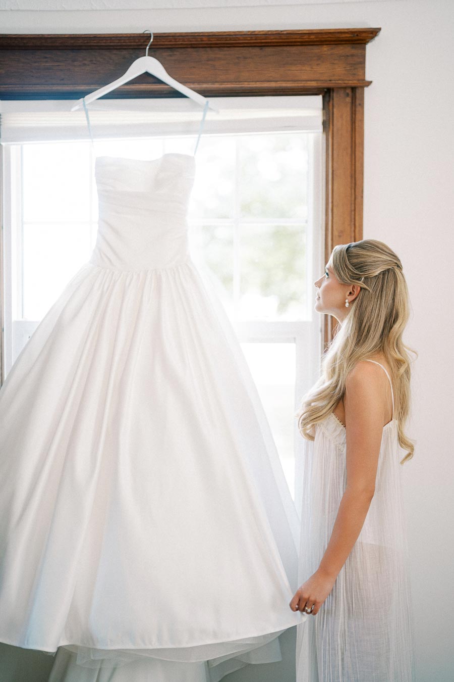 A bride standing beside her elegant white wedding dress hanging near a sunny window, preparing for her special day.