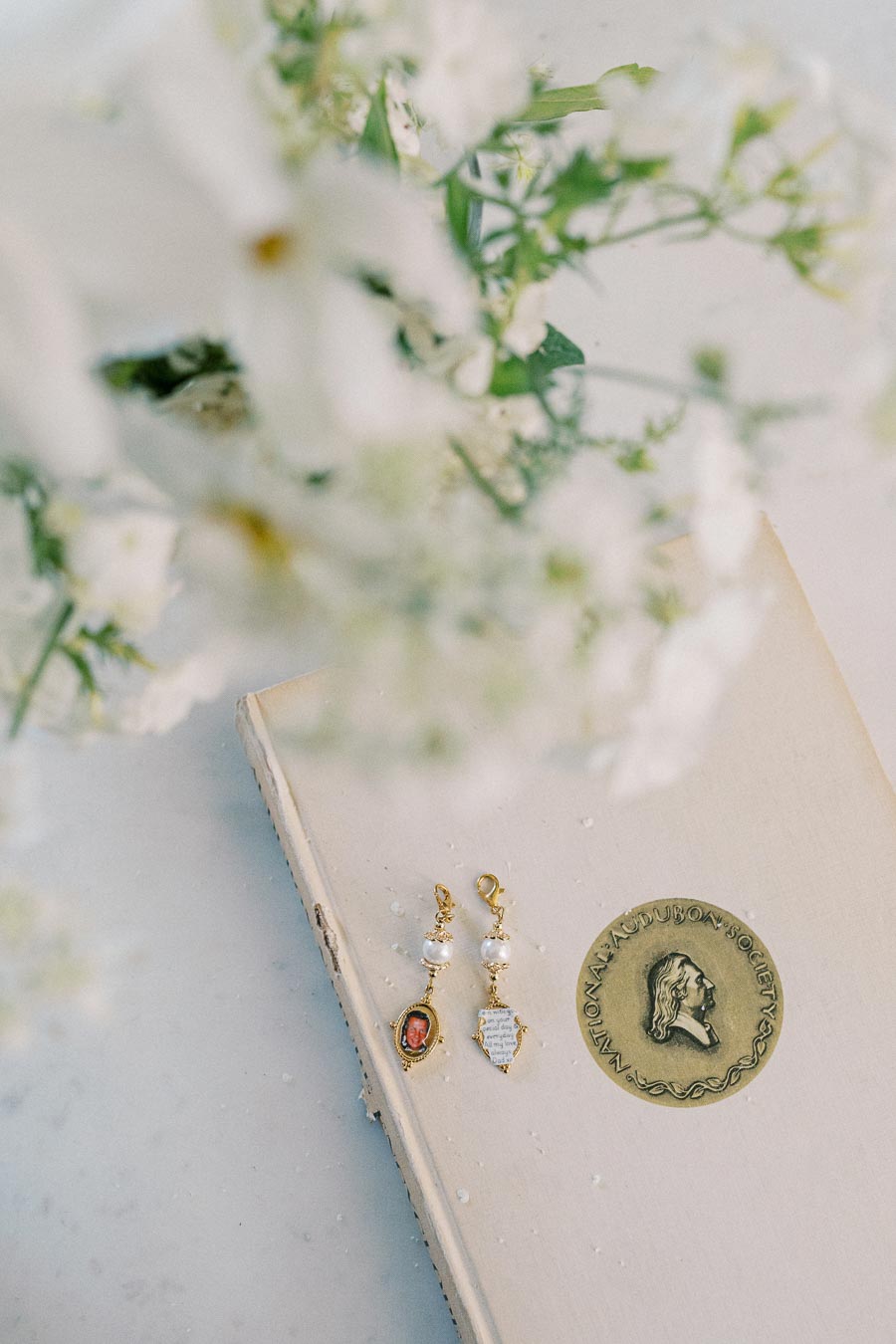 Delicate gold earrings with pearls placed on a vintage book, accompanied by soft-focus white flowers in the foreground.