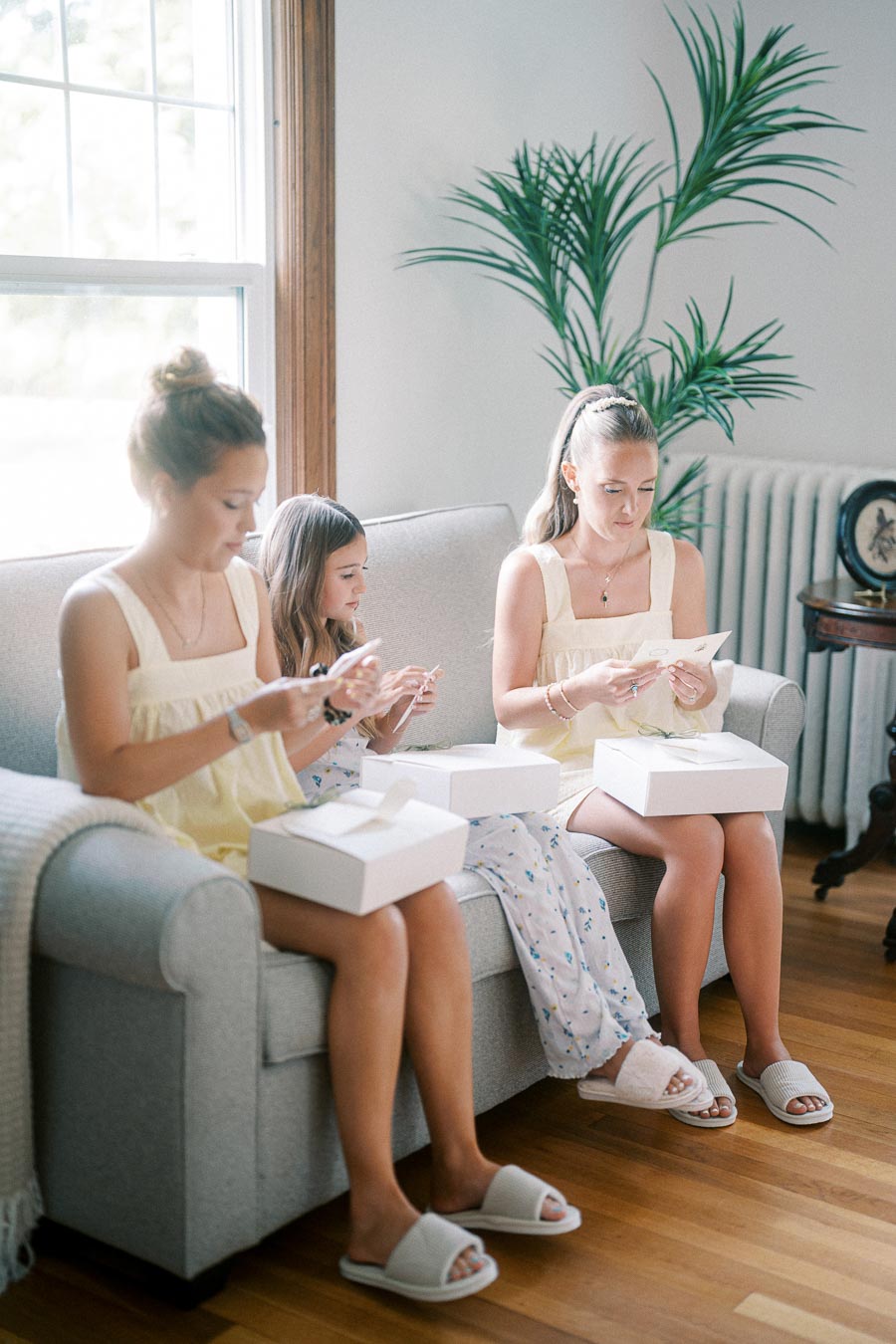 Three young women sitting on a couch, opening white gift boxes, surrounded by a cozy and bright living room setting with natural light and a potted plant in the background.