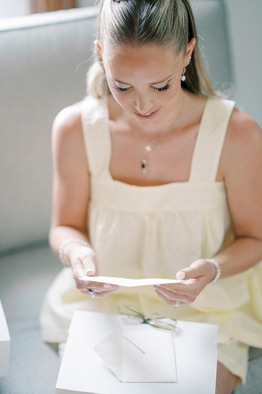 A woman in a light yellow dress reading a card while sitting on a sofa, with a white gift box and open envelope on her lap.