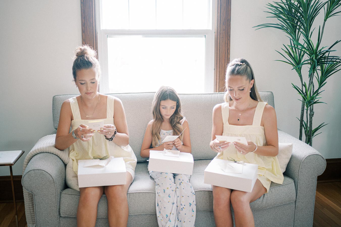 Three women sitting on a couch, each opening white gift boxes and reading cards, with a potted plant in the background. They appear to be enjoying a special occasion in a sunlit room.