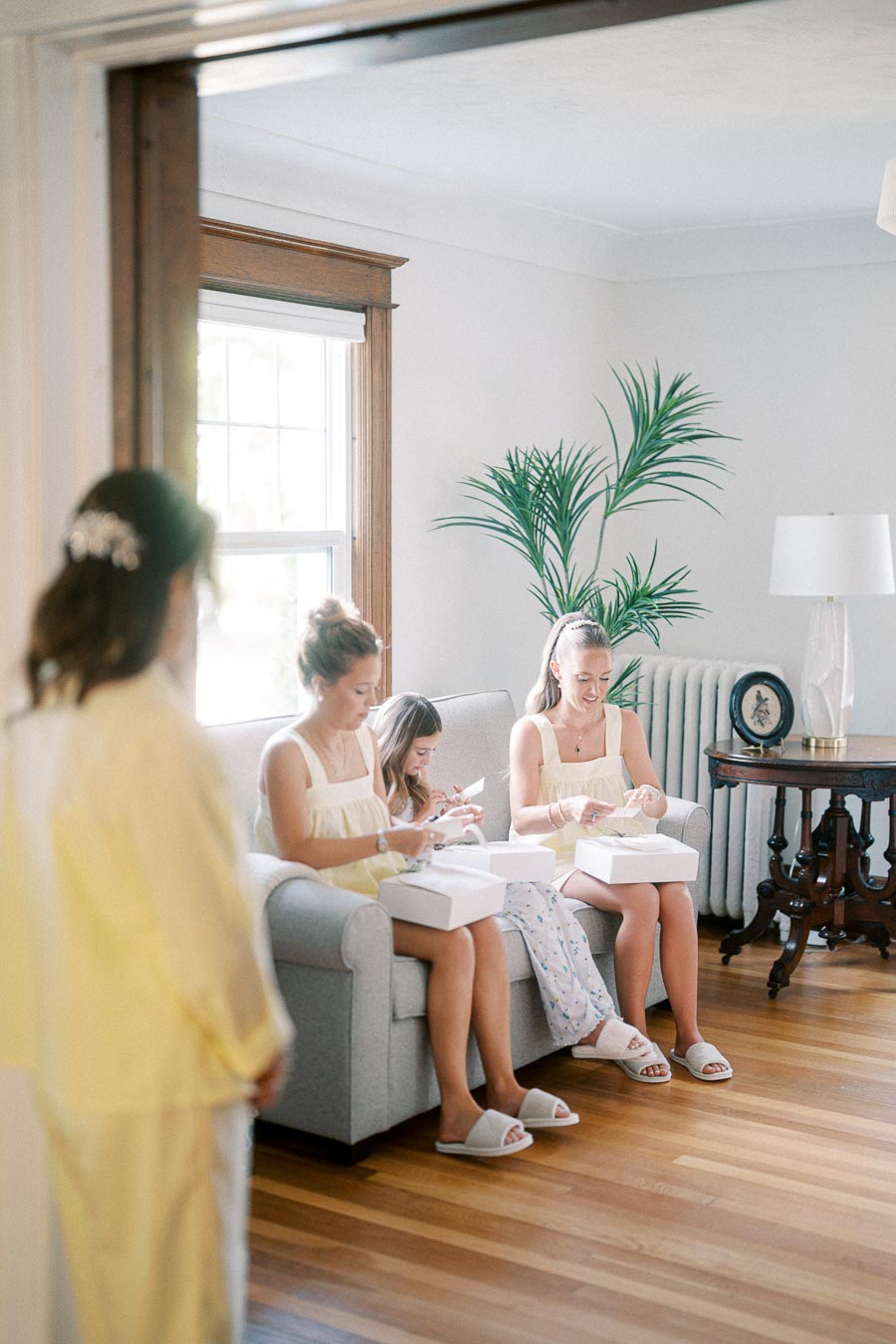 A group of young women in matching yellow dresses sitting on a sofa, opening white gift boxes in a bright living room with hardwood floors and a large window, adjacent to a standing woman in focus wearing similar attire.