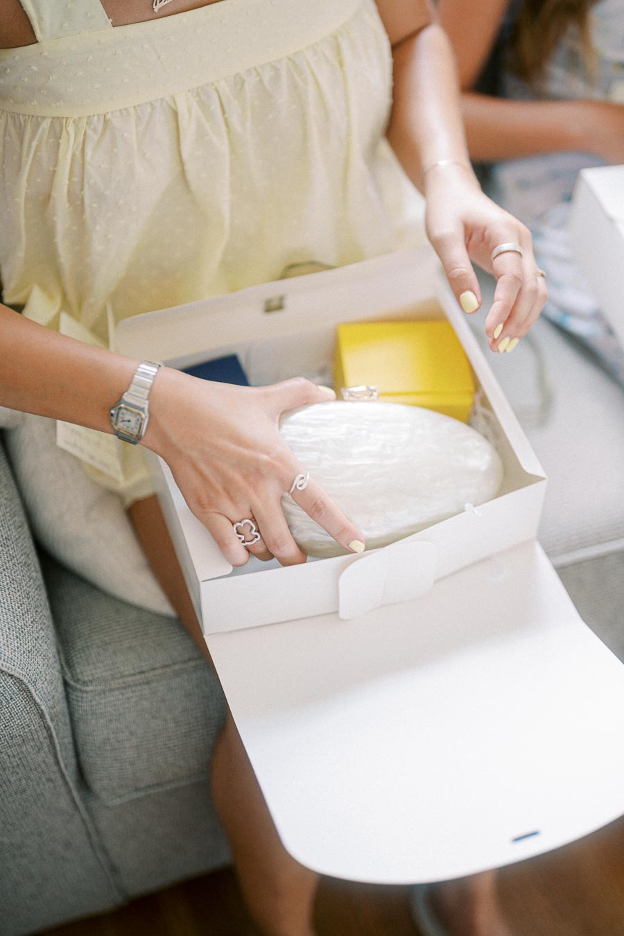 Woman in a light yellow dress opening a white gift box containing a packaged round item, with colorful boxes inside, sitting on a grey couch.