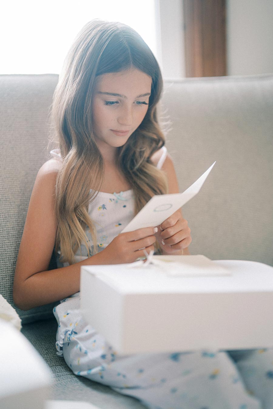 A young girl with long hair sits on a couch, reading a card with a white gift box in front of her. She wears a floral dress and appears thoughtful.