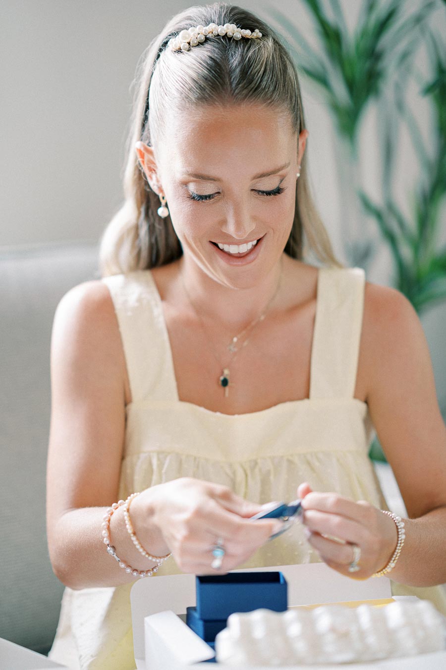 A smiling woman in a yellow dress opens a blue gift box, adorned with pearl jewelry, sitting on a comfortable couch with potted plants in the background.