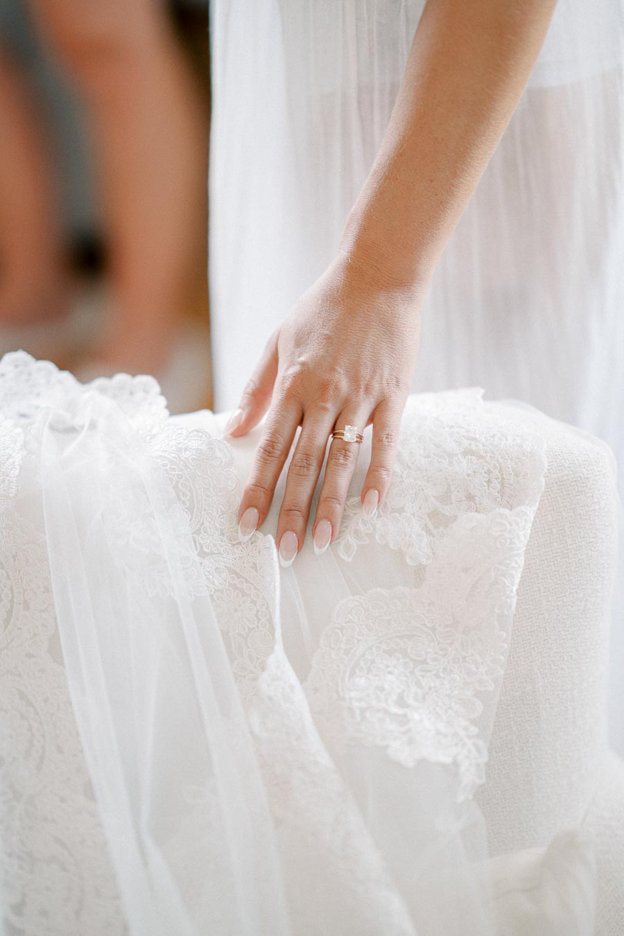 A close-up of a woman's hand gently resting on a delicate white lace wedding dress, showcasing a sparkling engagement ring.