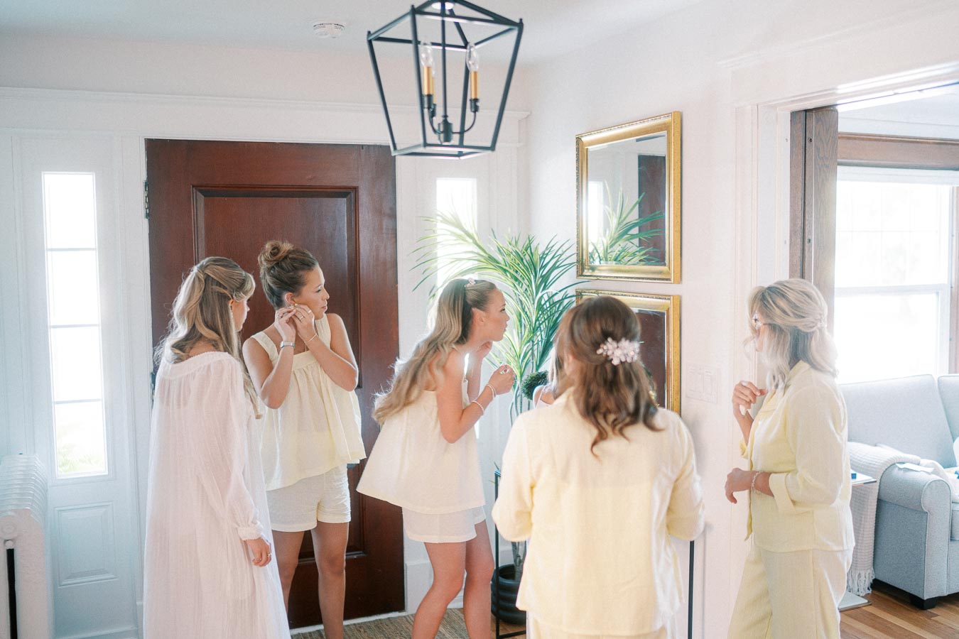 A group of women in light, casual attire getting ready together in a bright, elegant living room. They are adjusting their jewelry and hair, while conversing and smiling in front of large mirrors adorned with gold frames. The room is well-lit with natural light, featuring a wooden door, indoor plants, and modern lighting fixtures.