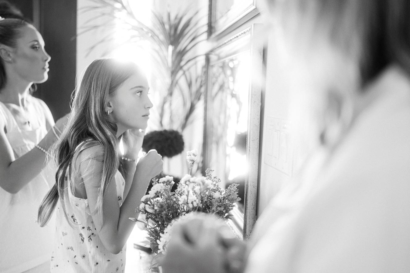 A young girl with long hair and a floral dress looks at herself in a mirror, adjusting her necklace, with sunlight streaming in from the window. A woman stands beside her, also looking into the mirror, in a softly lit room with decorative plants.