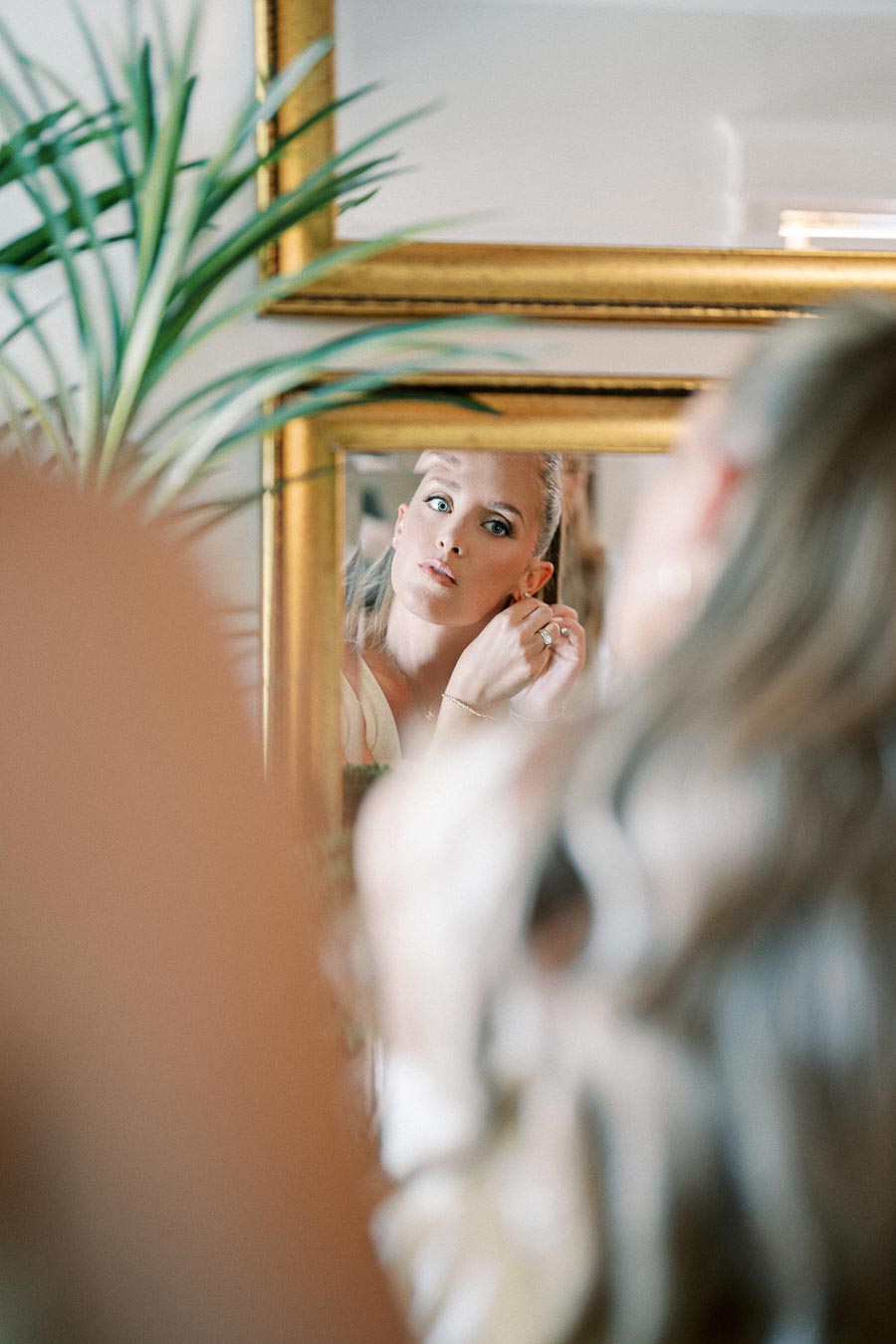 Woman adjusting earrings in front of a mirror with a gold frame, reflecting her focused expression, surrounded by soft lighting and blurred foreground elements.