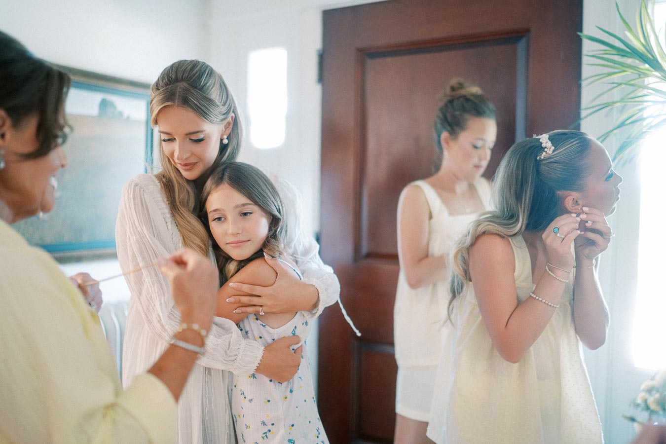 A bride embraces a young girl while surrounded by bridesmaids getting ready in a cozy room, capturing a warm and intimate moment before the wedding ceremony. The scene includes women adjusting their jewelry, conveying a sense of joy and anticipation.
